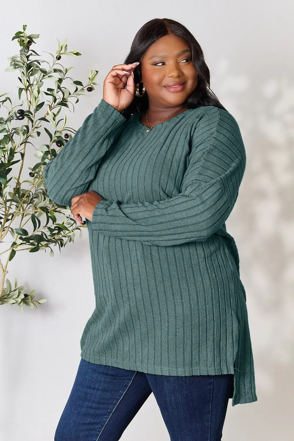 Woman in green ribbed long sleeve sweater and jeans, standing by leafy plant