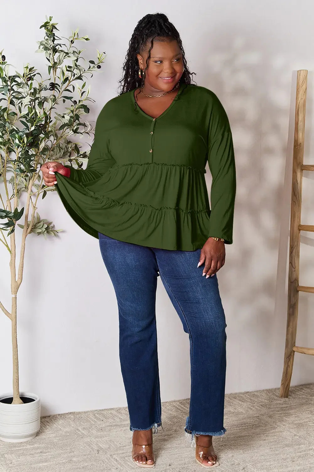Smiling woman in long sleeve green tiered blouse and blue jeans, indoor casual fashion