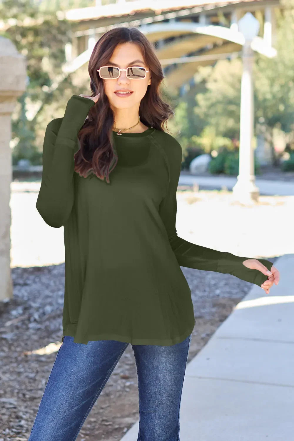 Woman in olive green long sleeve top and blue jeans posing outdoors in sunglasses