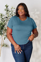 Woman wearing a blue short sleeve t-shirt and jeans, posing indoors by a leafy plant.