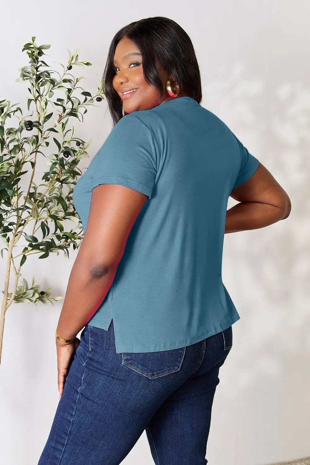 Woman modeling a teal short sleeve t-shirt and dark blue jeans, standing indoors near a plant