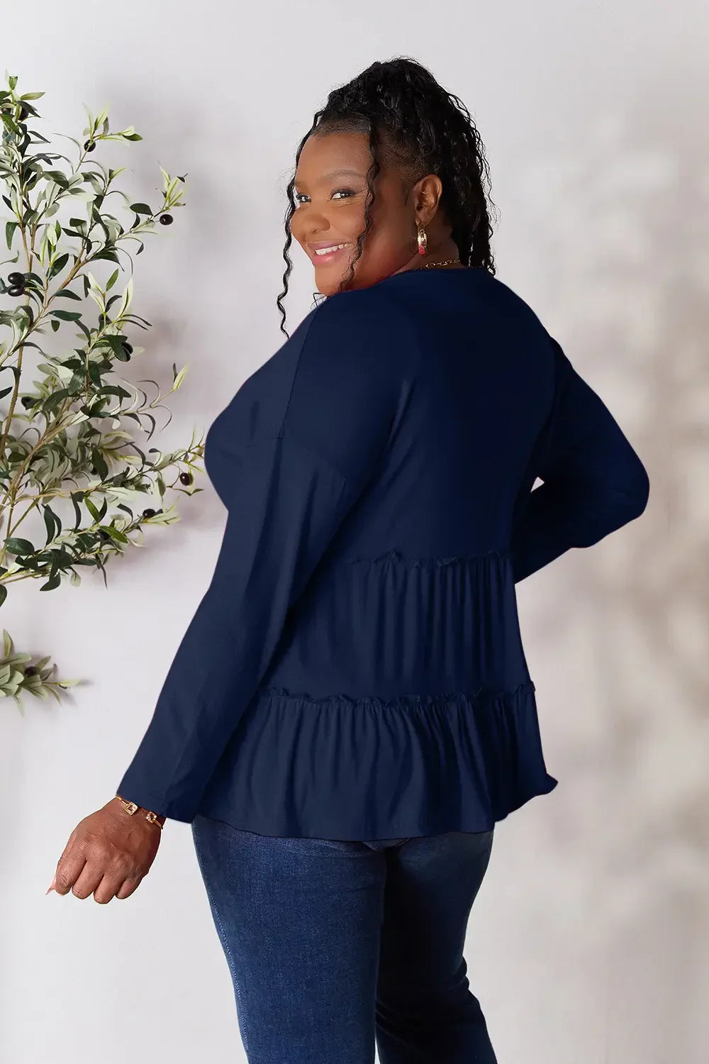 Smiling woman in a navy long-sleeve ruffle top and blue jeans standing by a plant
