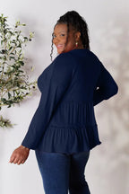 Smiling woman in a navy long-sleeve ruffle top and blue jeans standing by a plant