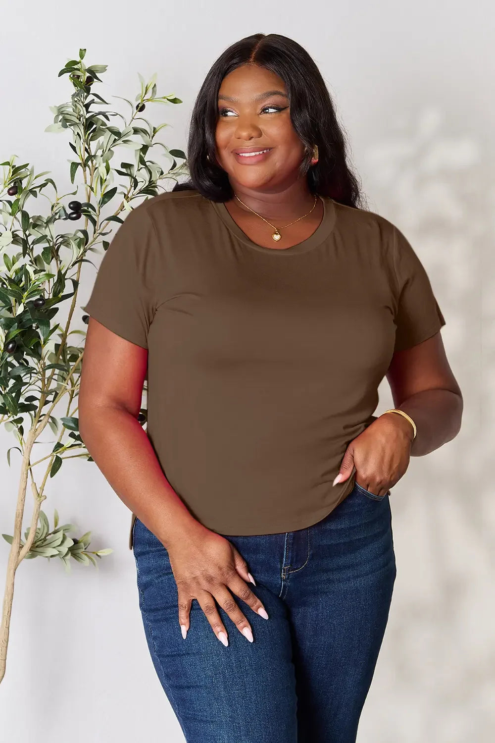 Woman wearing brown short sleeve t-shirt and blue jeans, standing by a plant