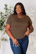 Woman wearing brown short sleeve t-shirt and blue jeans, standing by a plant