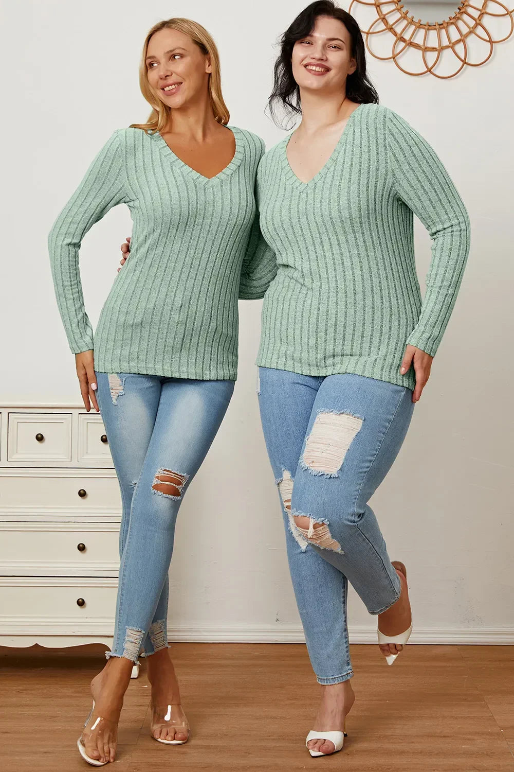 Two women in mint green ribbed sweaters and ripped blue skinny jeans posing indoors