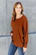 Woman in a brown long sleeve top and blue jeans standing against a concrete wall