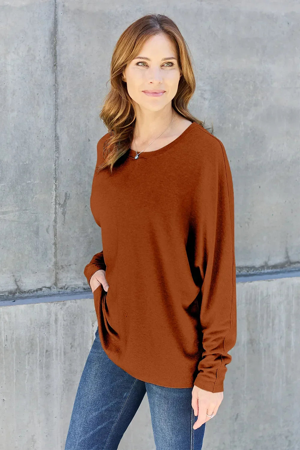 Woman in a brown long sleeve top and blue jeans standing against a concrete wall