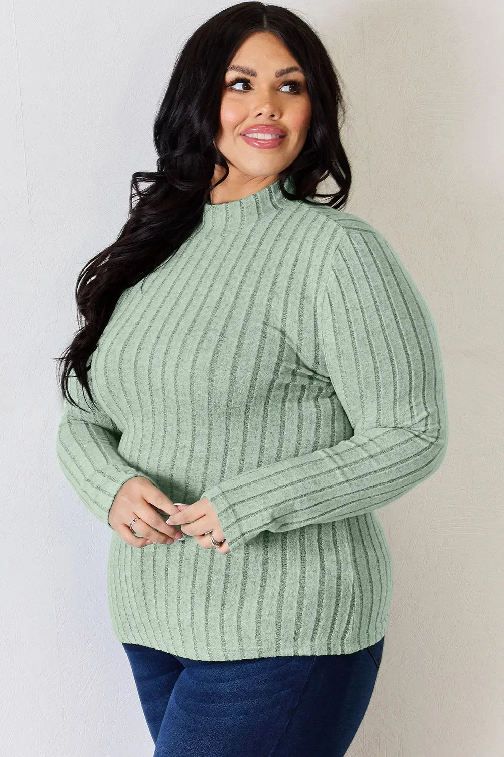 Woman wearing a light green ribbed knit sweater and blue jeans, posing indoors.