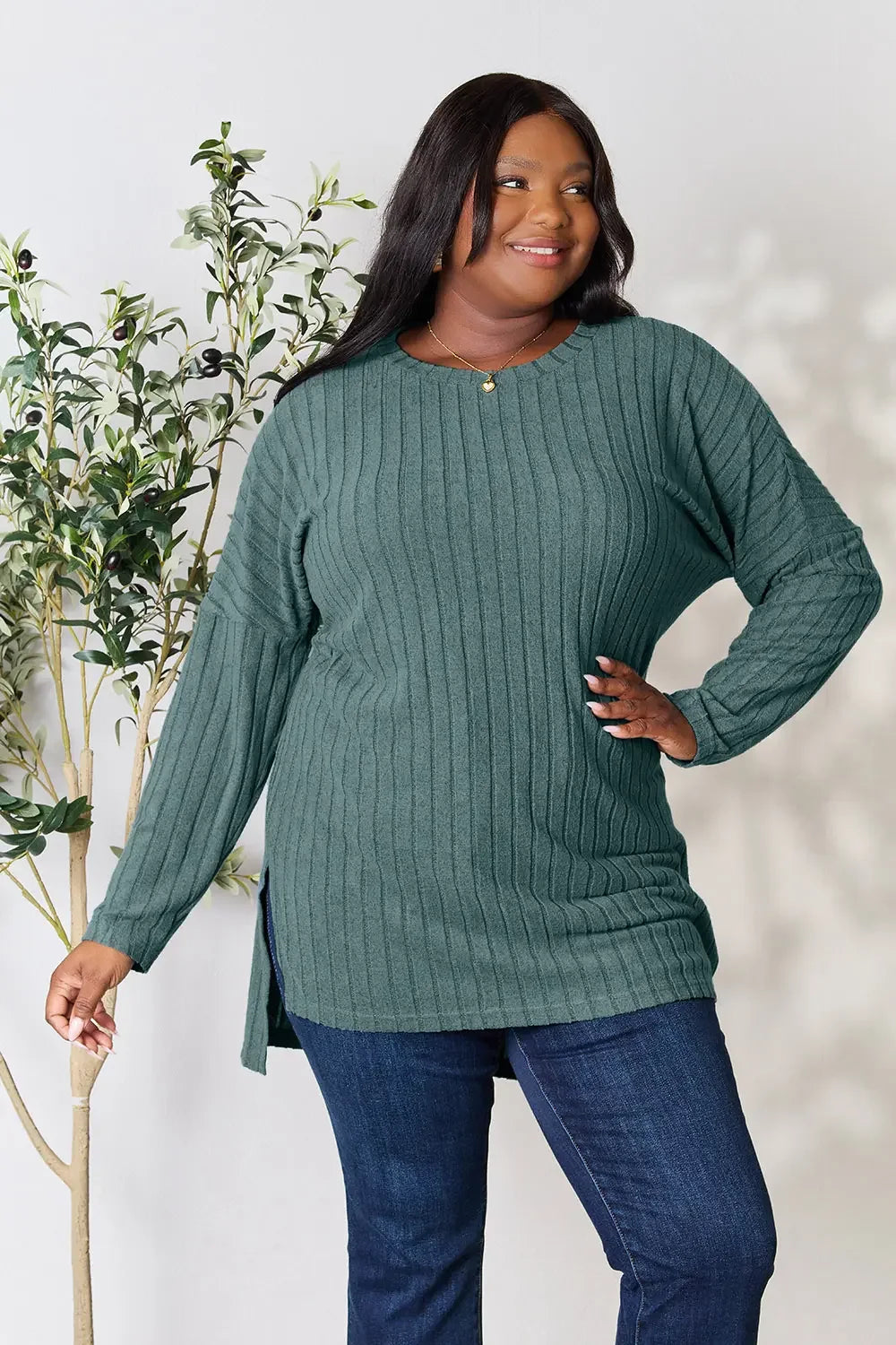 Woman wearing a ribbed green sweater and blue jeans, standing near a leafy plant