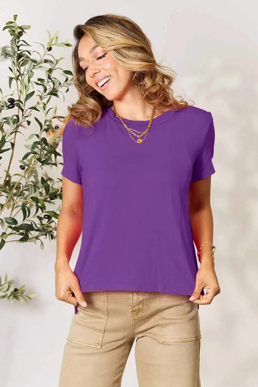 Woman wearing a purple short sleeve t-shirt and beige pants, smiling indoors.