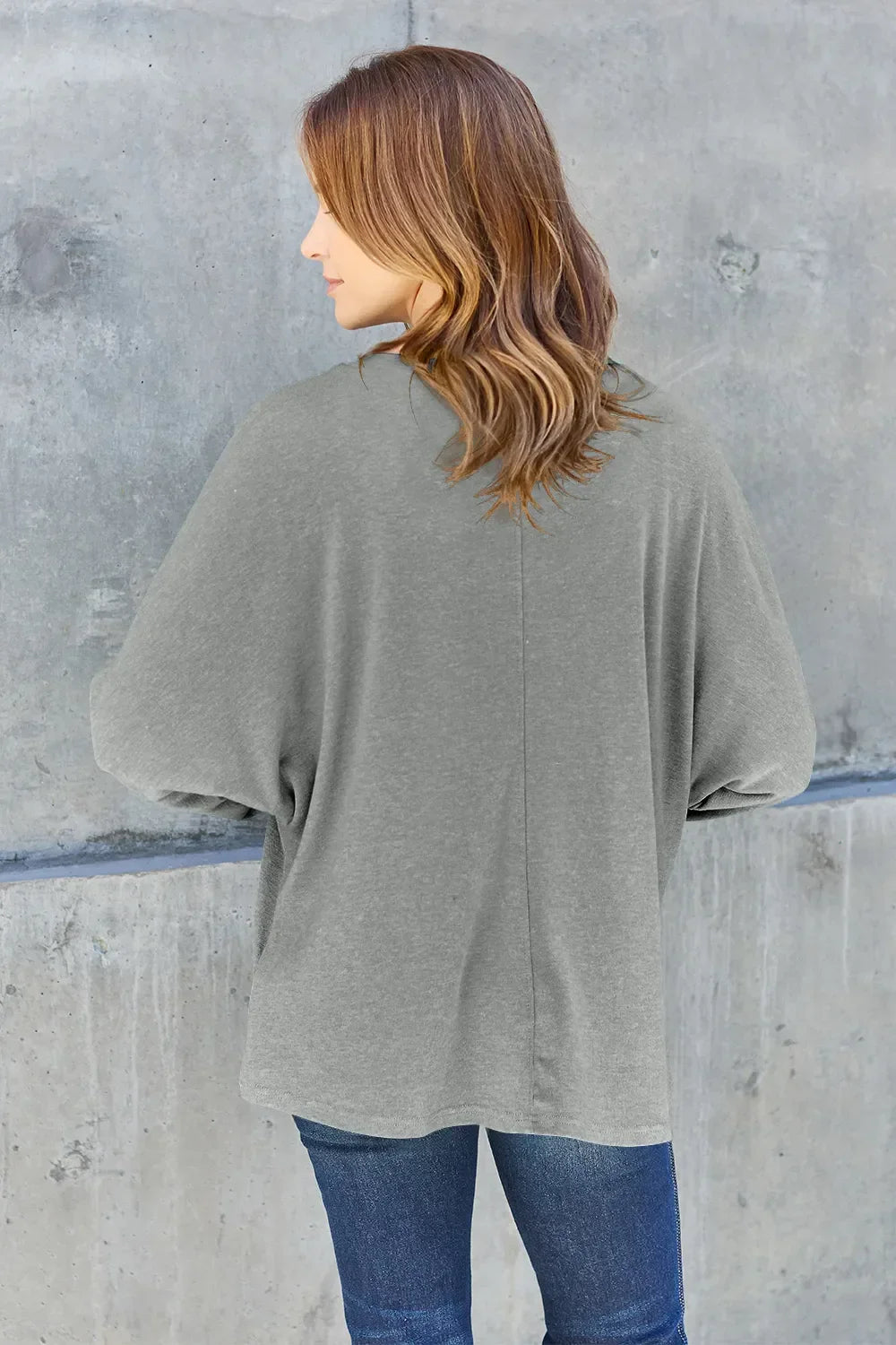Woman in casual grey long sleeve top and blue jeans standing against concrete wall