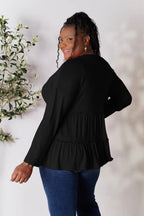 Woman wearing a black long sleeve blouse with ruffle details and blue jeans, smiling indoors