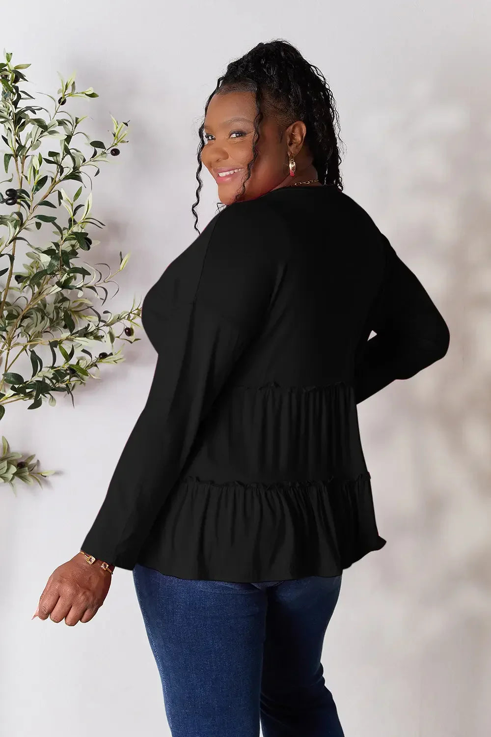 Woman wearing a black long sleeve blouse with ruffle details and blue jeans, smiling indoors