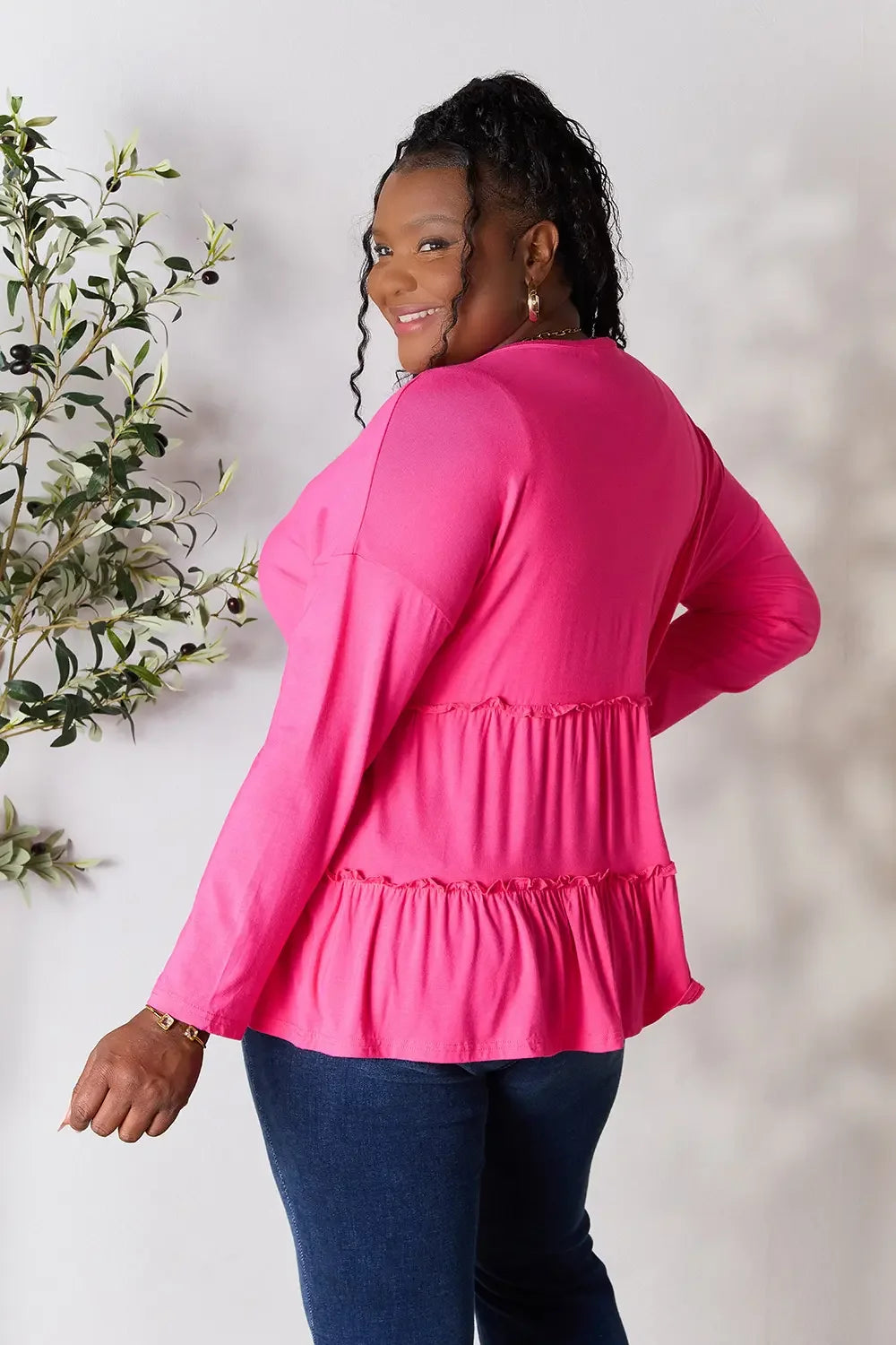 Smiling woman wearing a long sleeve pink ruffle top and blue jeans, standing sideways indoors.