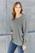 Woman wearing a casual grey long sleeve top with blue jeans, standing against a concrete wall.