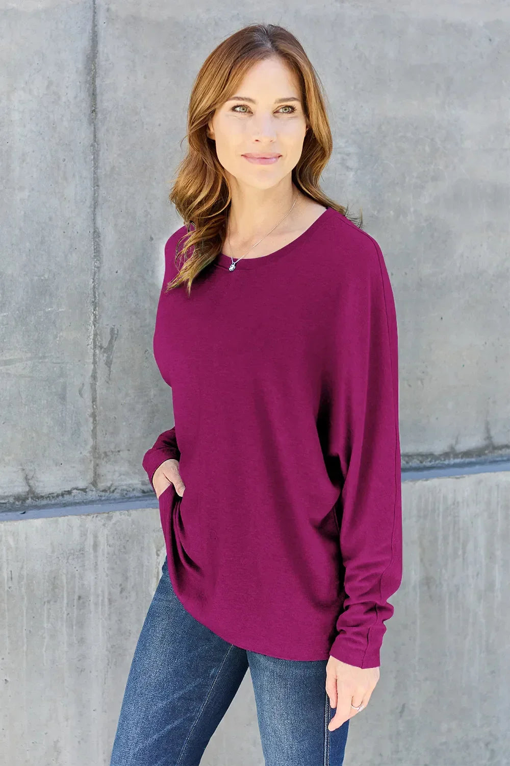 Woman wearing a long sleeve purple top and blue jeans standing against a concrete wall