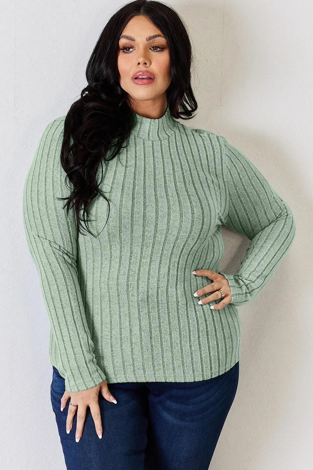 Woman wearing light green ribbed knit sweater and dark blue jeans against a light wall