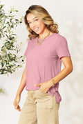 Woman in pink short sleeve t-shirt and beige pants, smiling, with gold jewelry, indoor setting.