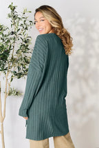Woman in teal ribbed long sweater and beige pants, standing by leafy plant, smiling