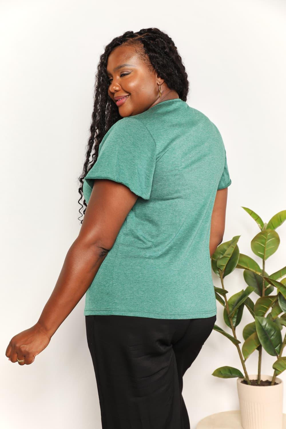 Woman modeling a green short-sleeve top and black pants, standing by a potted plant