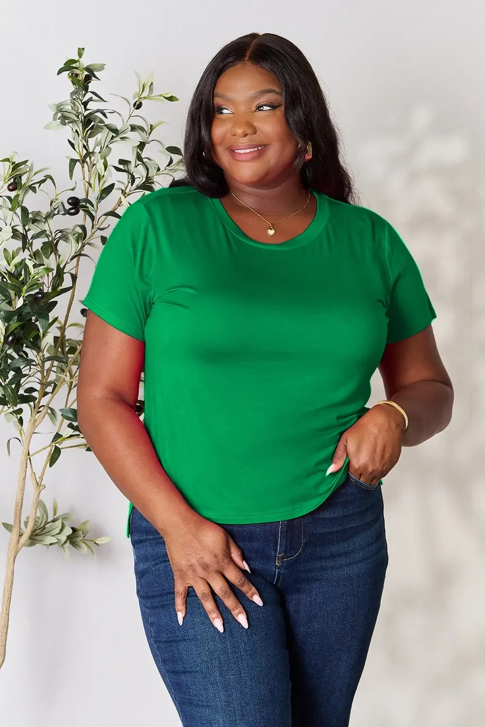 Woman in green short sleeve t-shirt and blue jeans, posing indoors by a plant