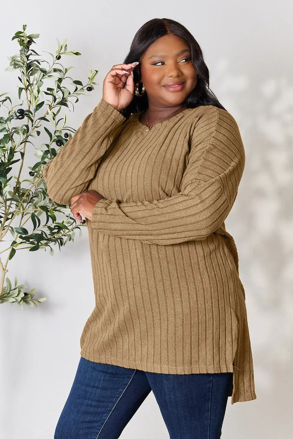 Woman wearing a ribbed tan knit sweater with long sleeves and blue jeans, standing indoors.