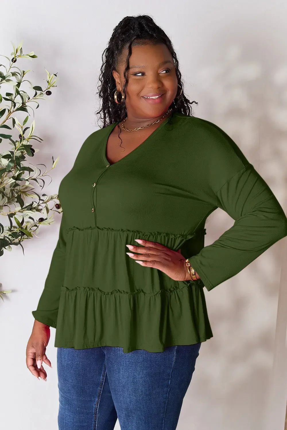 Woman wearing an olive green tiered long sleeve top and blue jeans, smiling indoors.