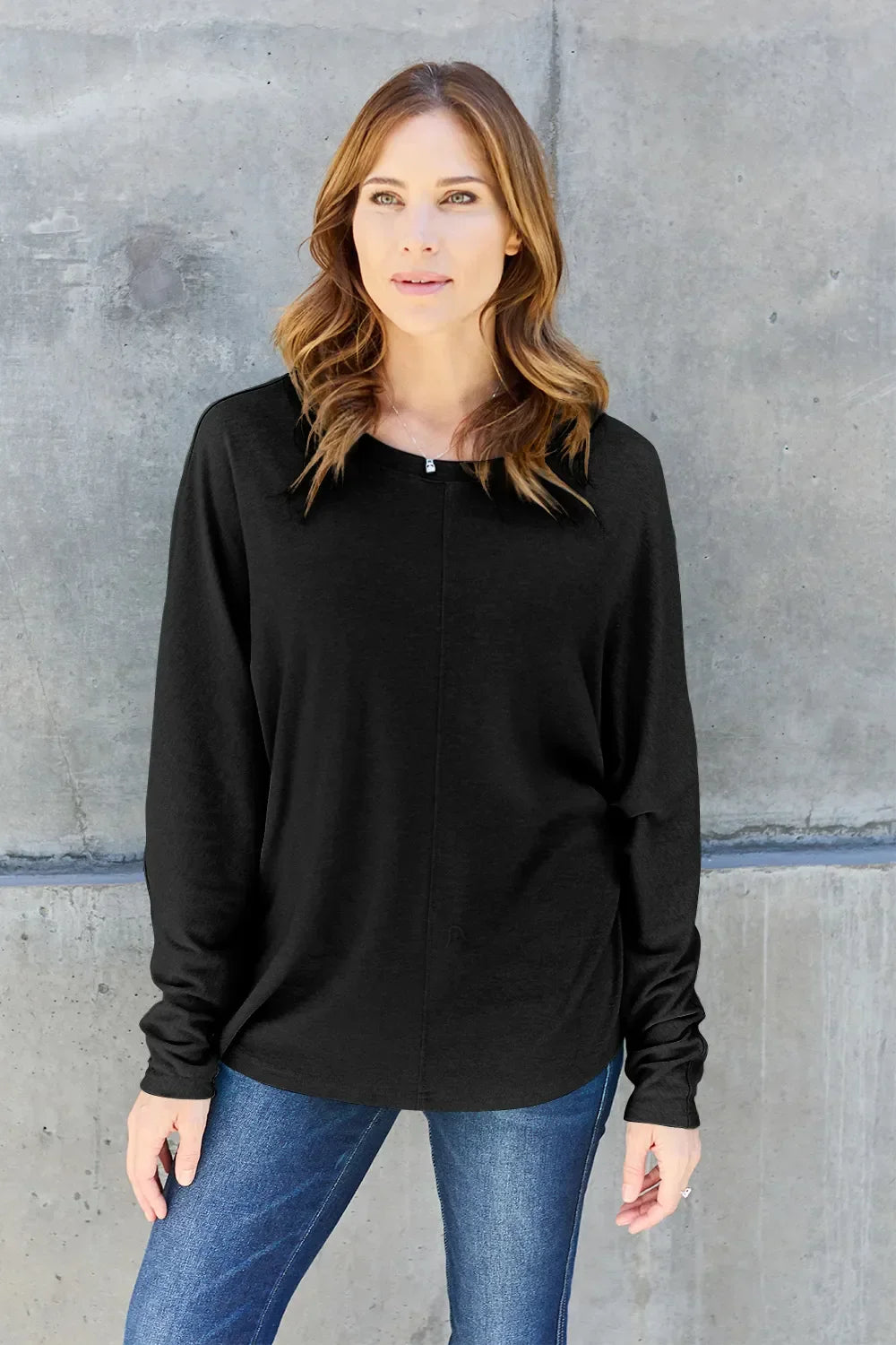 Woman wearing a black long sleeve top and blue jeans standing by a concrete wall
