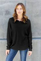 Woman wearing a black long sleeve top and blue jeans standing by a concrete wall