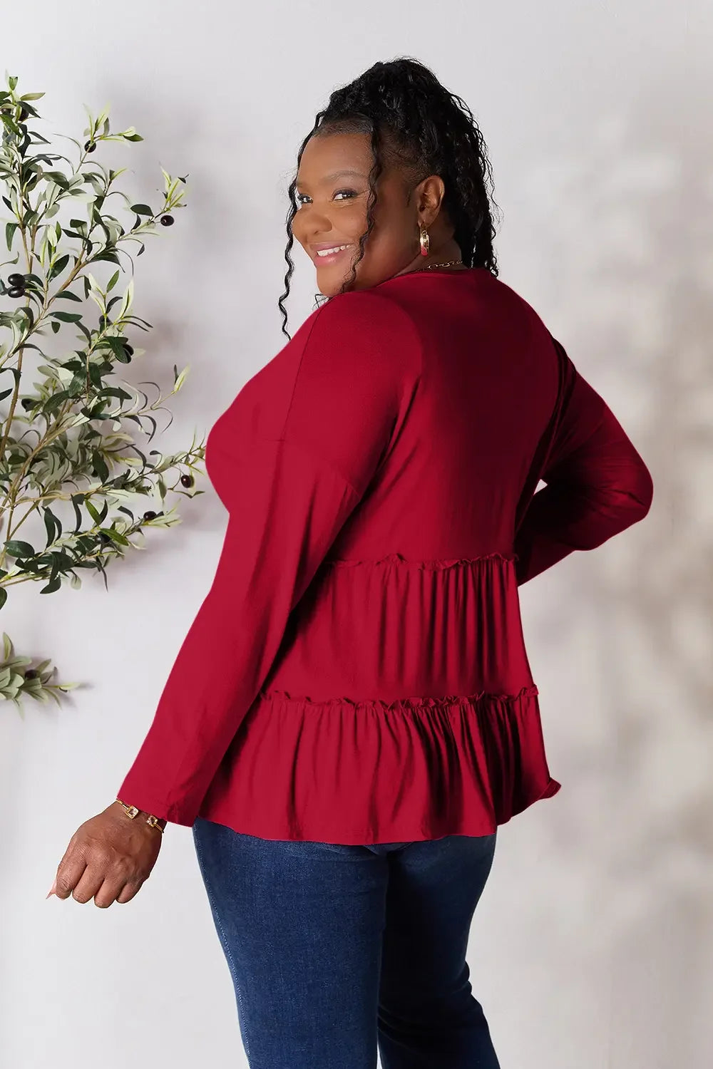 Smiling woman in long sleeve red ruffle blouse and blue jeans, standing by leafy plant