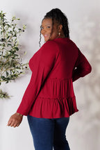 Smiling woman in long sleeve red ruffle blouse and blue jeans, standing by leafy plant