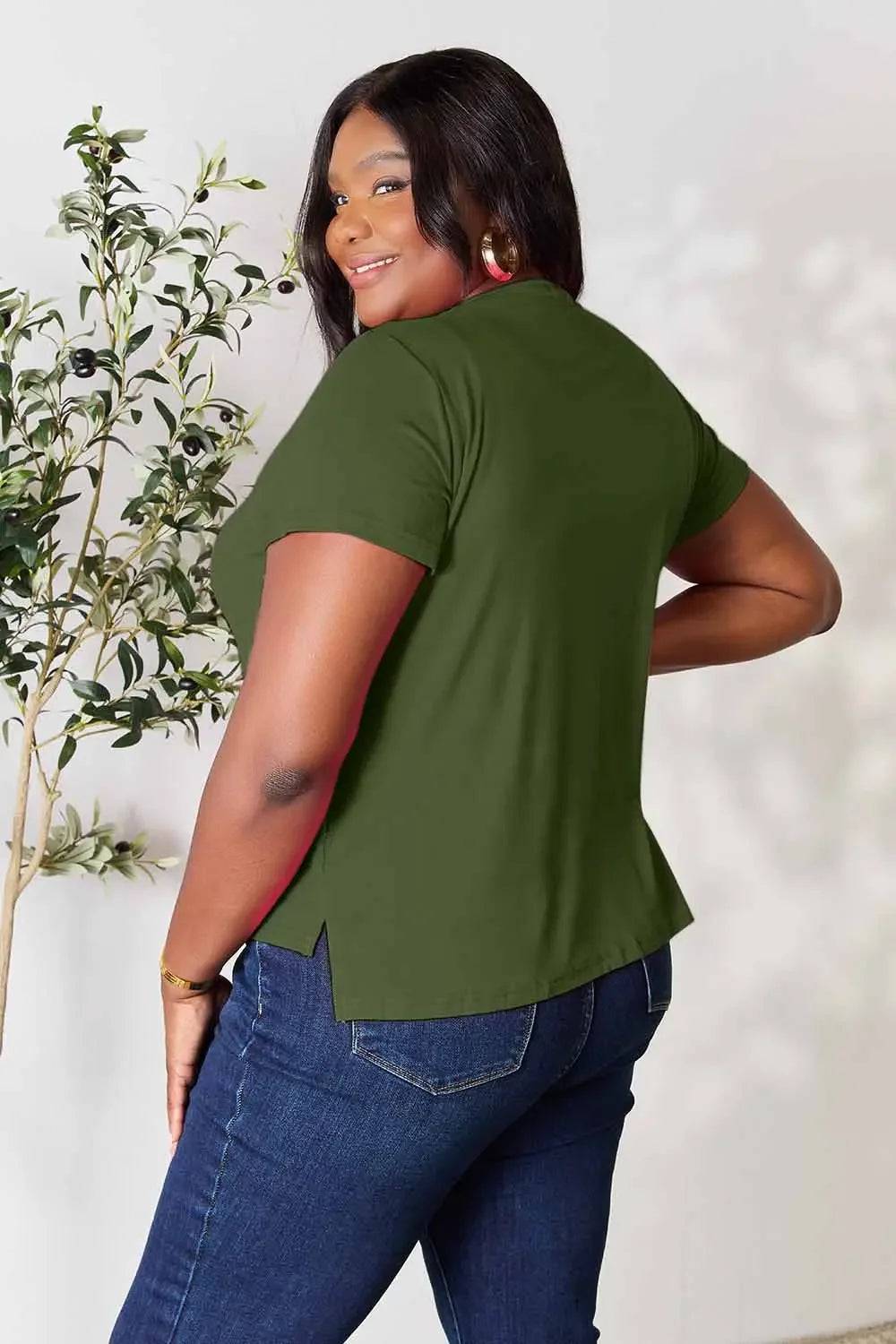 Woman in olive green short sleeve top and blue jeans, posing indoors near a leafy plant.