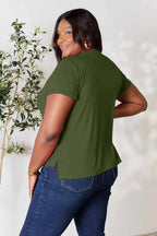 Woman in olive green short sleeve top and blue jeans, posing indoors near a leafy plant.