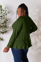 Woman in a long sleeve olive green ruffle blouse and blue jeans, smiling indoors