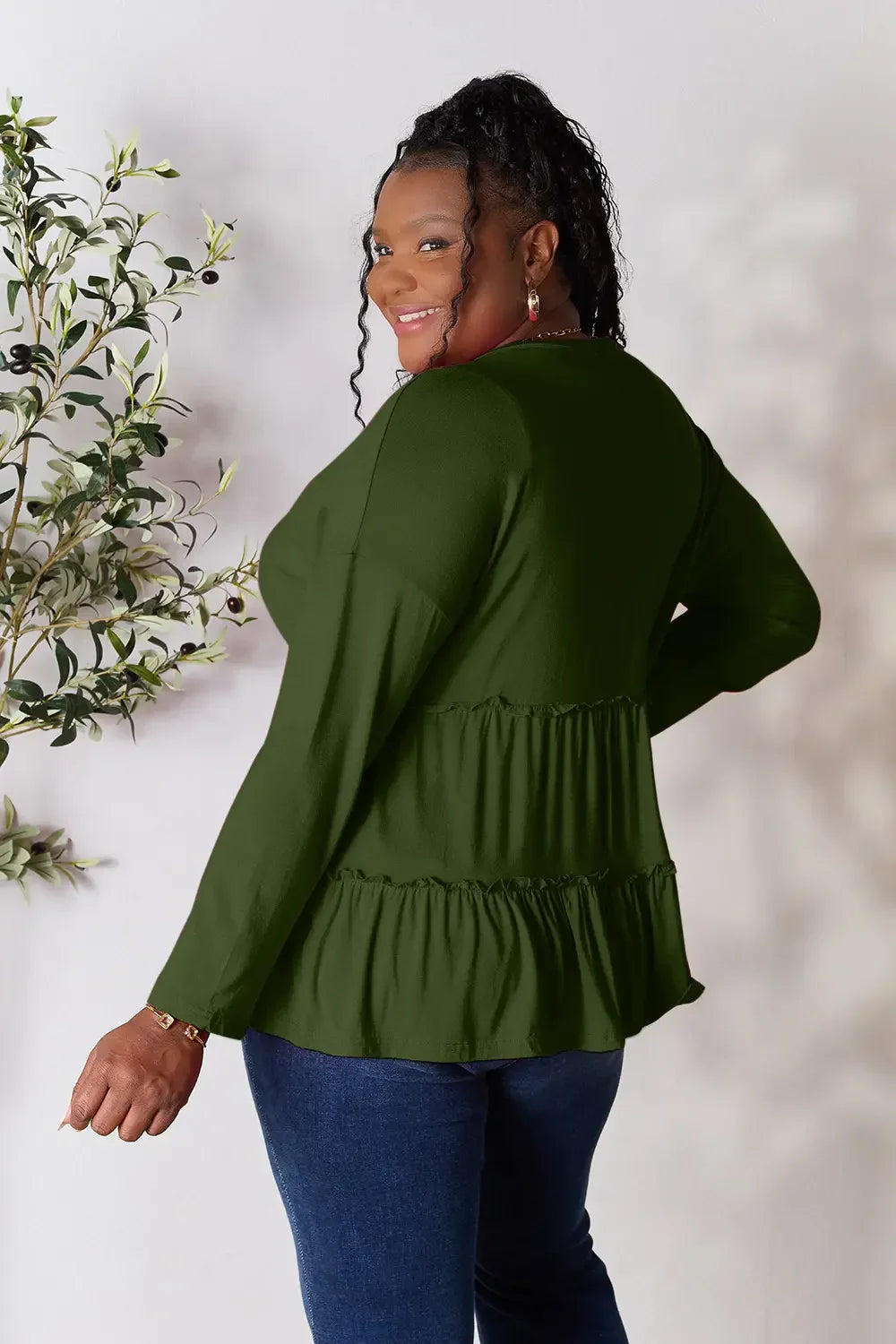 Woman in a long sleeve olive green ruffle blouse and blue jeans, smiling indoors