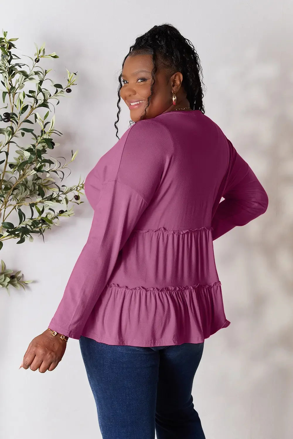 Smiling woman wearing a ruffled long sleeve magenta top and dark blue jeans, standing indoors