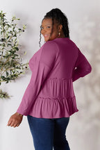 Smiling woman wearing a ruffled long sleeve magenta top and dark blue jeans, standing indoors