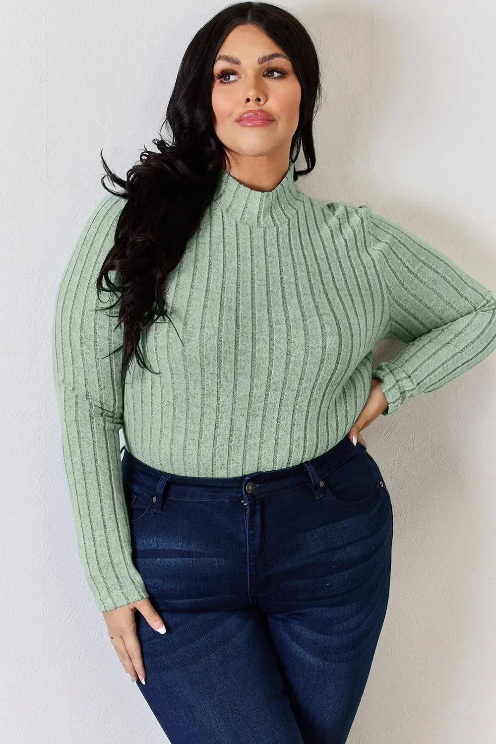 Woman wearing light green ribbed knit sweater and dark blue jeans, posing against a white wall