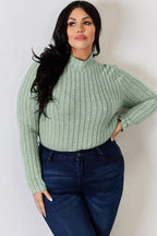 Woman wearing light green ribbed knit sweater and dark blue jeans, posing against a white wall
