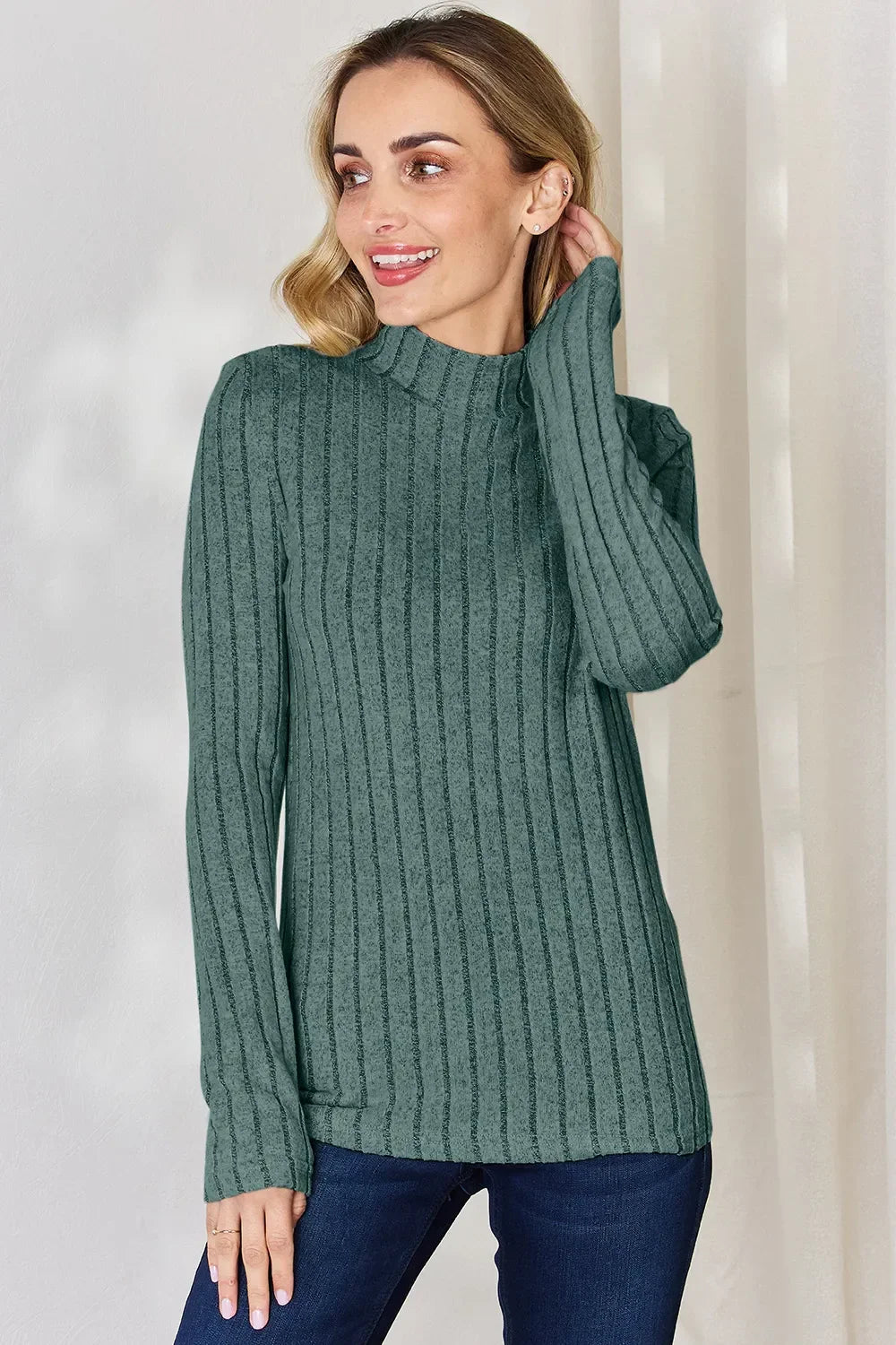 Woman in green ribbed knit long sleeve top and blue jeans standing indoors