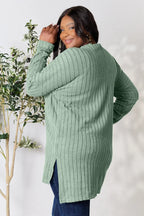 Woman in a sage green ribbed knit sweater and dark jeans, posing indoors by a plant.