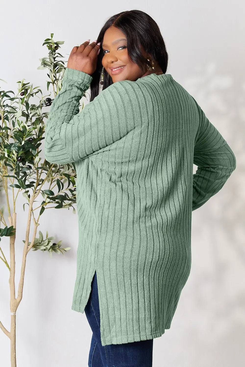 Woman in a sage green ribbed knit sweater and dark jeans, posing indoors by a plant.