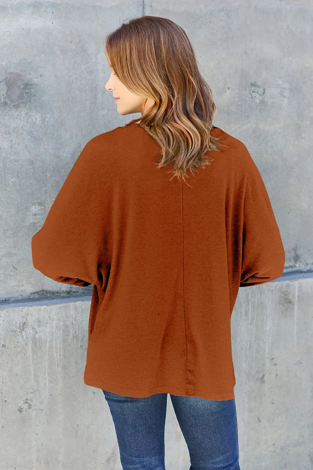 Woman in loose brown sweater with rolled sleeves and denim jeans, standing by concrete wall