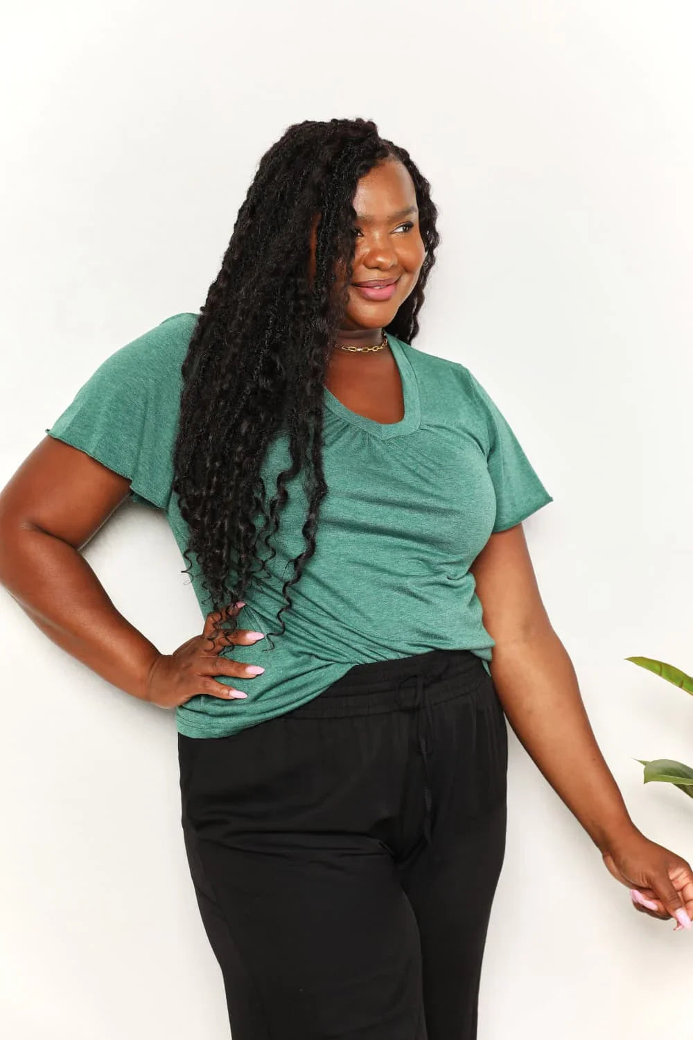 Woman wearing a green short sleeve V-neck top and black pants, posing against white background
