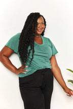 Woman wearing a green short sleeve V-neck top and black pants, posing against white background