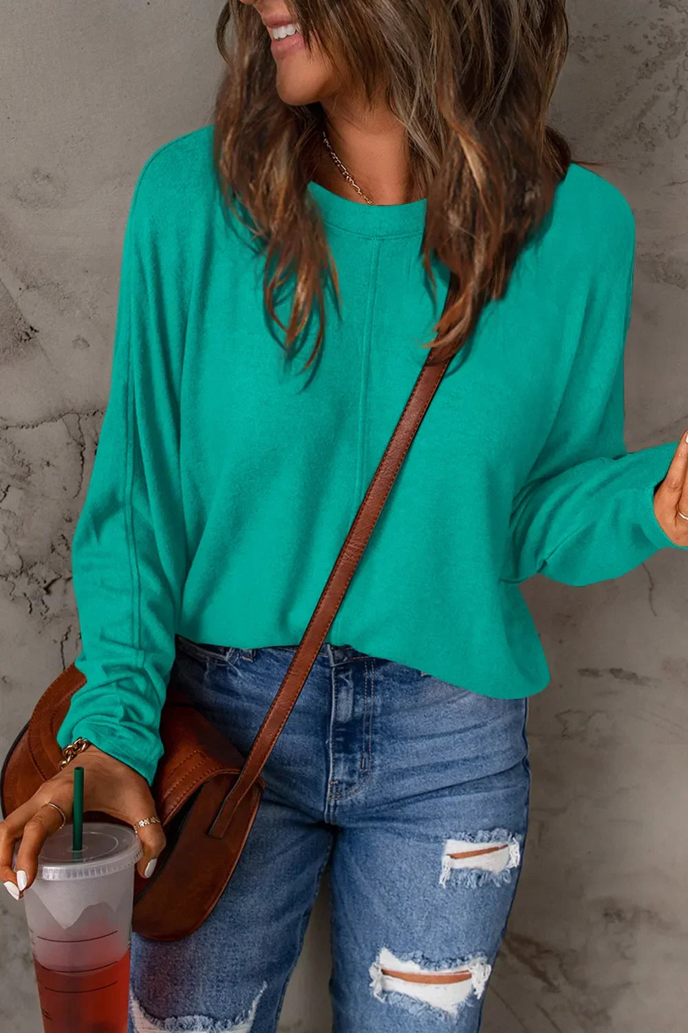 Woman in teal long sleeve top and ripped blue jeans holding a drink, casual street style