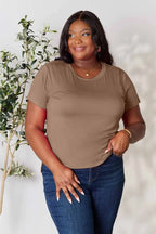 Woman wearing a brown short-sleeve t-shirt and blue jeans, posing indoors by a plant