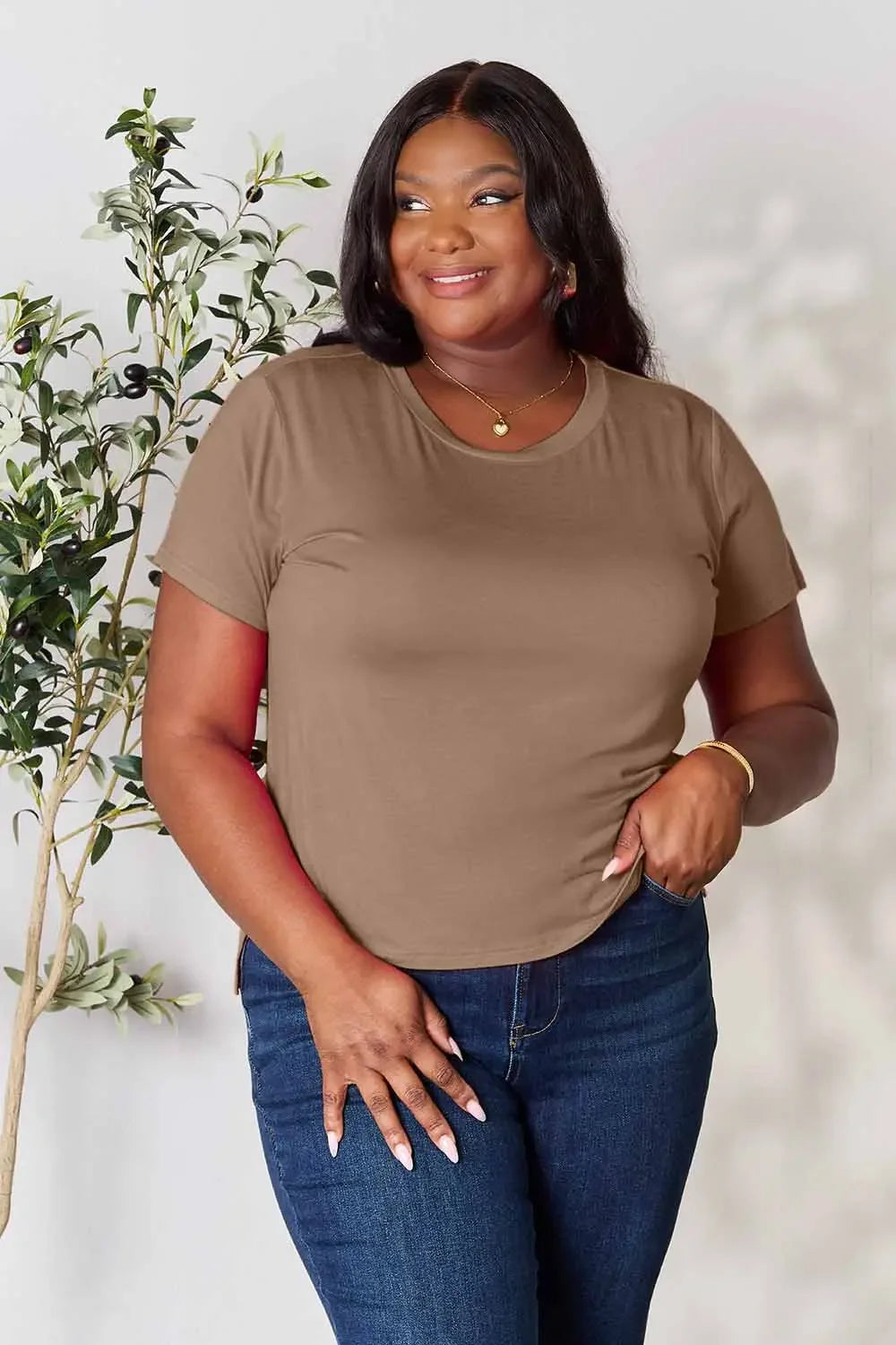 Woman wearing a brown short-sleeve t-shirt and blue jeans, posing indoors by a plant
