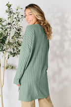 Woman wearing a ribbed sage green long cardigan sweater, standing near an olive tree.